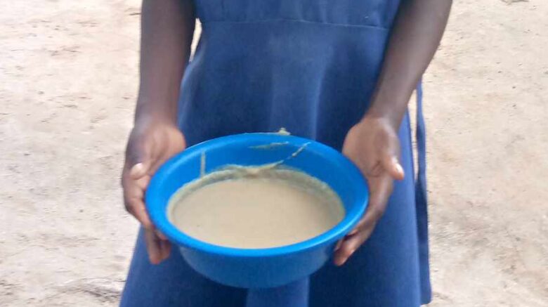 A girl in a blue school dress holds a small plastic bowl of porridge, in a sandy courtyard.
